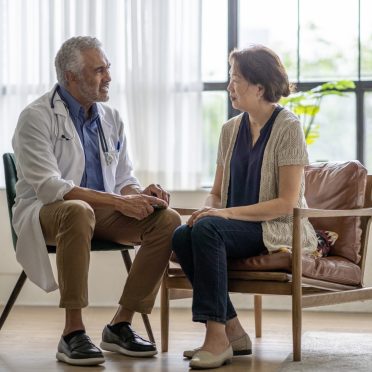 A male doctor of African decent sits with a senior patient in the comfort of her own home as he discusses with her some options for home care.  He is wearing a white lab coat and seated beside the patient who is dressed casually and listening attentively to the doctors suggestions.