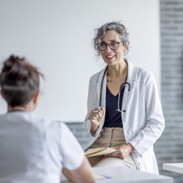 A mature female doctor sits at the front of a classroom as she teaches a small group of students.  She has a file folder on her lap as she talks with the students, and is dressed in a white lab coat.  The students are seated in front of her and paying close attention as they take notes.